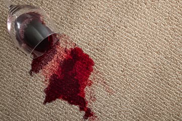 A tipped wine glass beside a fresh red wine spill on beige carpet.